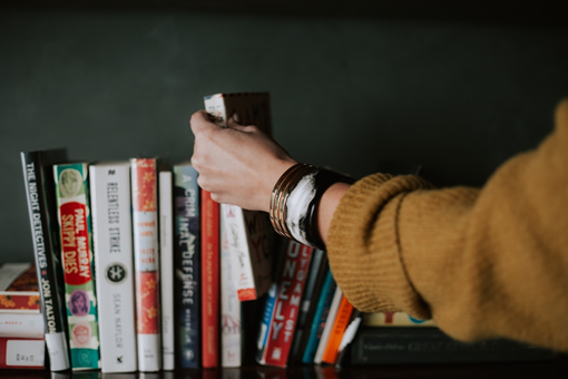 A woman's hand with an autumn sweater picking out a one of the cosiest autumn reads from a shelf.