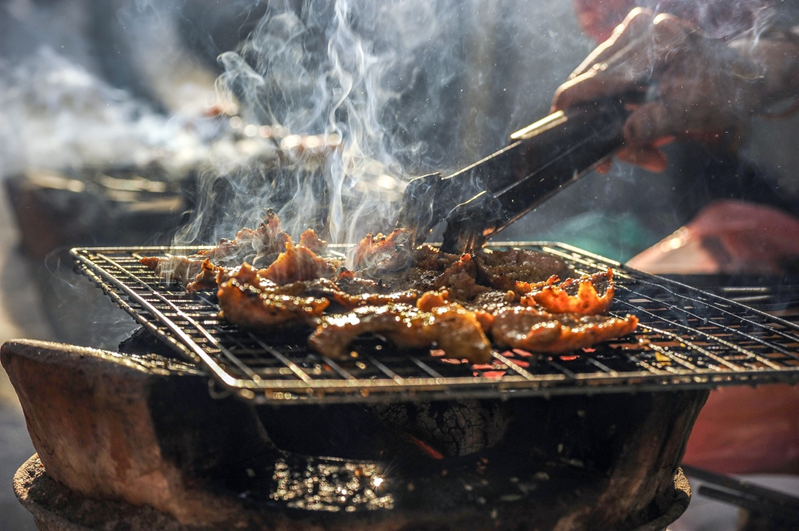 Ribs barbecuing on an open grill outside with a man's hand holding tongs over one of the meat cuts.