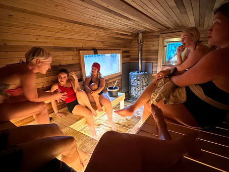 Five women in swimsuits relax and converse inside a wooden sauna. Warm lighting and steam create a calm, cozy atmosphere.