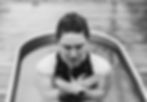 Person in a black swimsuit sits cross-armed in a cold plunge, eyes closed. Wood decking around. Calm and reflective mood.