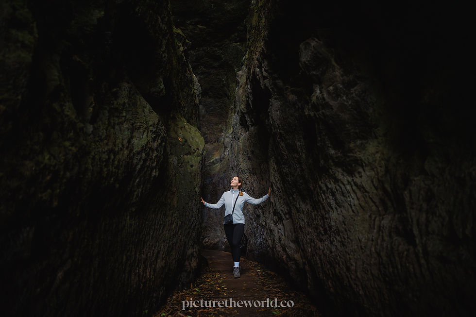 Person in a light jacket explores Levada do Furado (PR10) on Madeira a narrow, dark rocky crevice, touching walls. Mossy textures and dim lighting create a mysterious mood.