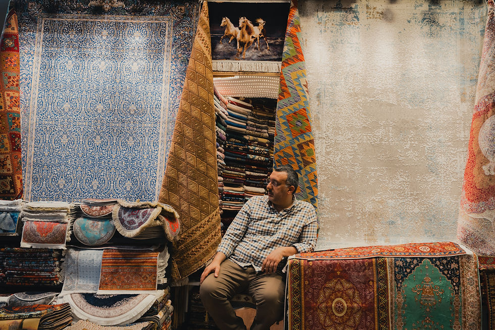Man sitting among colorful patterned rugs in Grand Baazar. Background features stacked textiles and a horse tapestry, creating a cozy atmosphere.