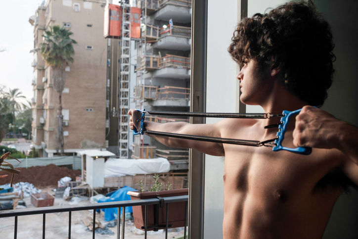 Saar stretching a chest expander by his home window toward a building under construction in Yad Eliyahu, documentary photography Flora Malinash.