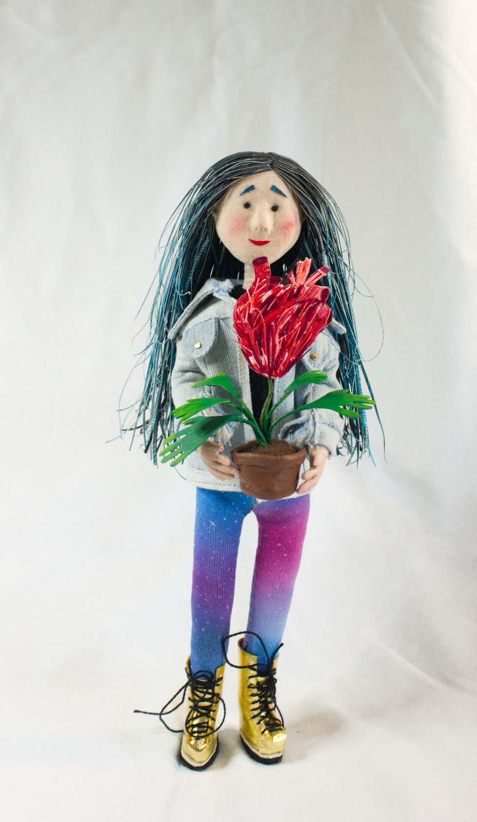 Handmade textile puppet of a young woman with long blue hair holding a red felt plant, created with fabric, felt, and embroidery details.