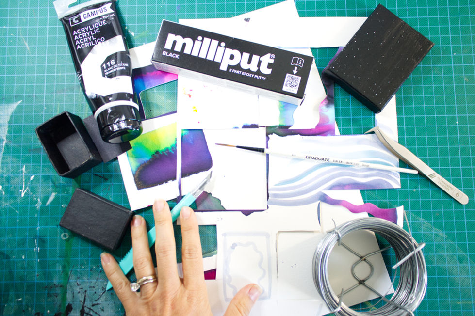 Artist’s hand over a green cutting mat with wires, Milliput, patterned paper, and tools for diorama making.
