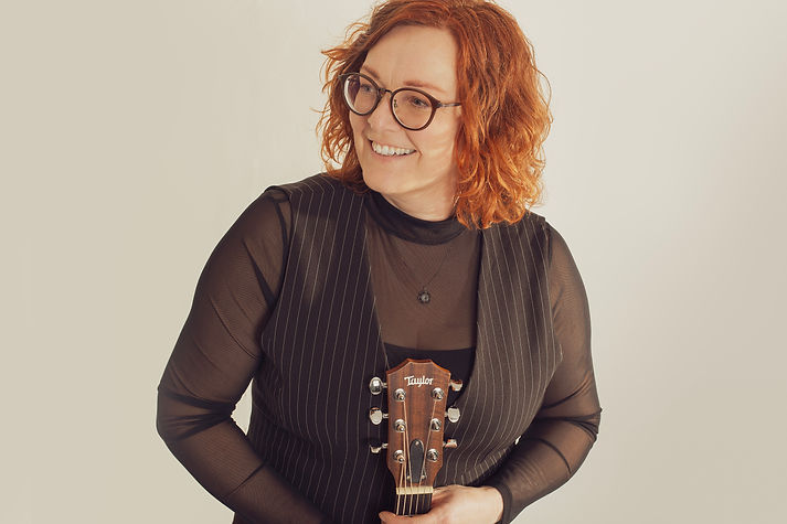 Smiling woman holding Taylor ukulele, wearing glasses, looks away, portrait, Anna Sellén StorySinger.