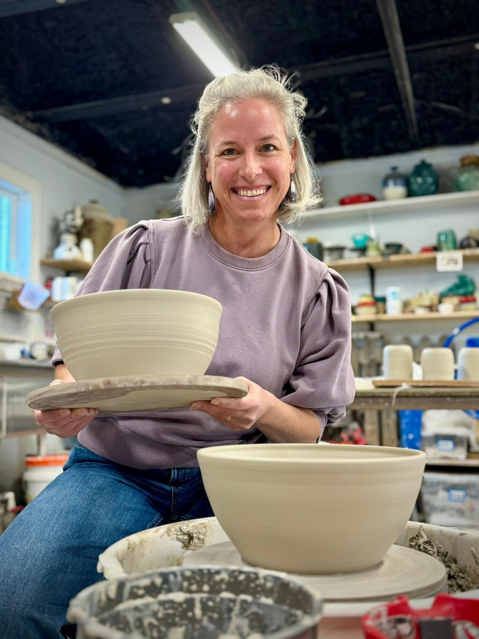 A person seated at a pottery wheel shapes a clay vessel with both hands, surrounded by studio tools and shelves of ceramic pieces.