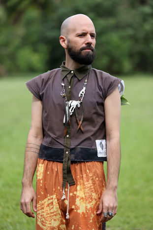 Man wearing an upcycled outfit with a brown sleeveless button up and orange printed pants and a necklace made from bones and shells. Fashion design by Miami artist Pangea Kali Virga.