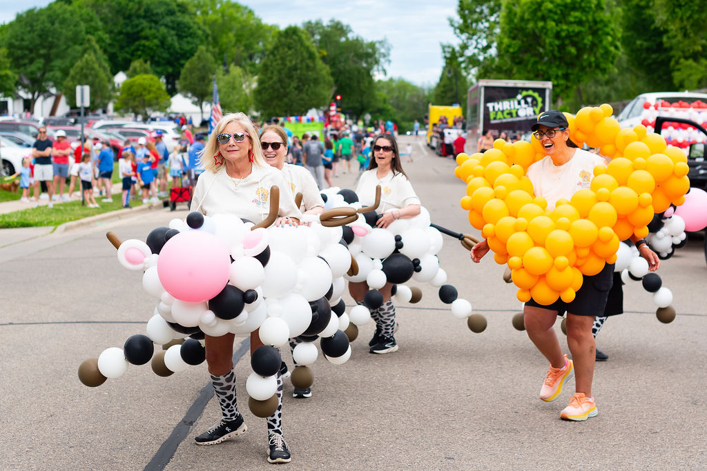 Walkers in balloon cow costumes and a yellow balloon cheese costume, marching in the Memorial Day parade.