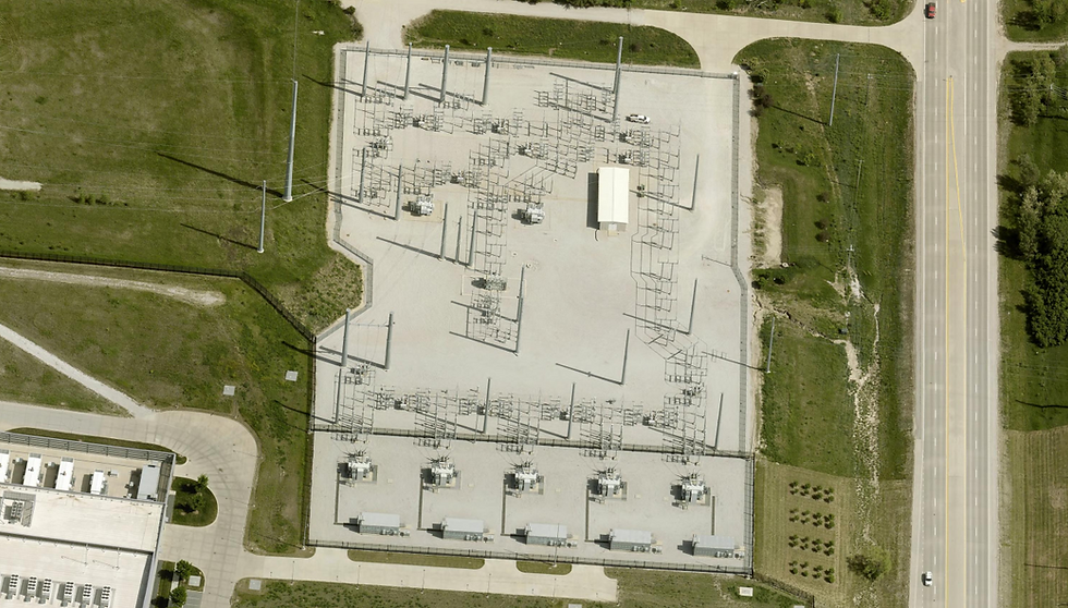Dedicated Transmission Substation, Springfield, Nebraska — adjacent to Meta Sarpy County Campus. This is where the abstraction of "power consumption" becomes concrete. The fenced compound occupying the center of the frame is a high-voltage transmission substation built exclusively to serve the data center visible at lower left. The large rectangular transformer banks arrayed along the southern perimeter step down incoming grid voltage — likely 115kV or 230kV transmission-level current — to the distribution voltage the campus can consume. The latticed steel structures throughout the yard are disconnect switches, bus bars, and surge arresters: the switching infrastructure that allows operators to isolate sections of the substation without interrupting load. The single small operations building at center is unstaffed under normal conditions; the entire facility runs on SCADA automation. High-voltage transmission lines enter from the east, visible as the tall steel poles along the right edge of the frame. What this image encodes is a singular fact: a single Meta campus in rural Nebraska requires dedicated transmission infrastructure that, in an earlier era, would have served an entire mid-sized city. Photo taken by May 8, 2025, source: MMCG Invest, LLC/EagleView