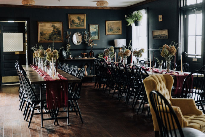2 long tables with black chairs. A black wall with landscape paintings and an oval mirror. Lots of natural light filters in through windows.