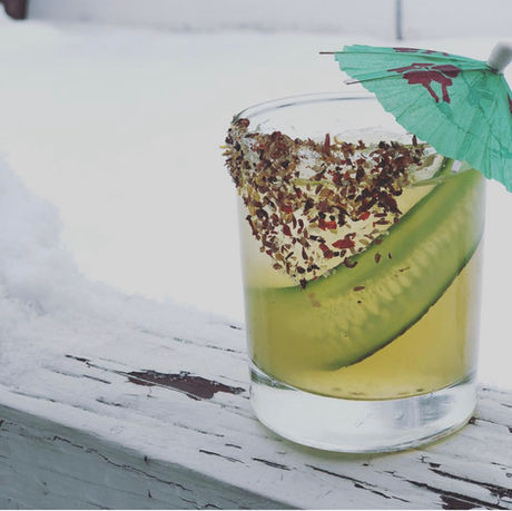 A pale green cocktail with a cucumber slice and smoked salt and a pale green drink umbrella sits on a snow covered white railing.
