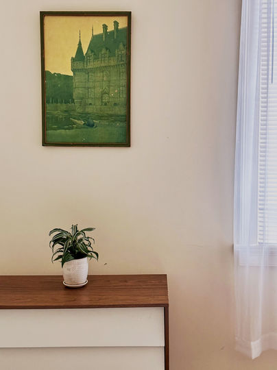 A white and wood toned dresser holds a small green plant. Above it is a painting of a castle using mostly green tones.