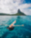 Uma turista flutua de braços abertos nas águas cristalinas da Praia da Conceição em Fernando de Noronha. Ela parece estar em total harmonia e relaxamento, cercada pelo vasto mar azul. Ao fundo, o imponente Morro do Pico se destaca, adicionando uma vista majestosa à cena. Esta imagem captura um momento de serenidade durante uma pausa em um passeio de canoa havaiana, um dos muitos atrativos oferecidos na ilha