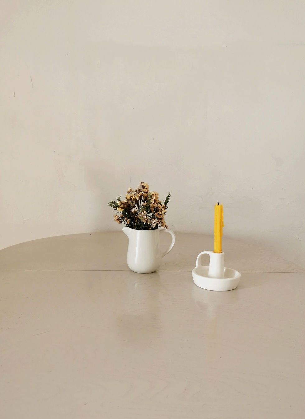 White pitcher with dry flowers, white ceramic candle holder with beeswax candle on white kitchen table