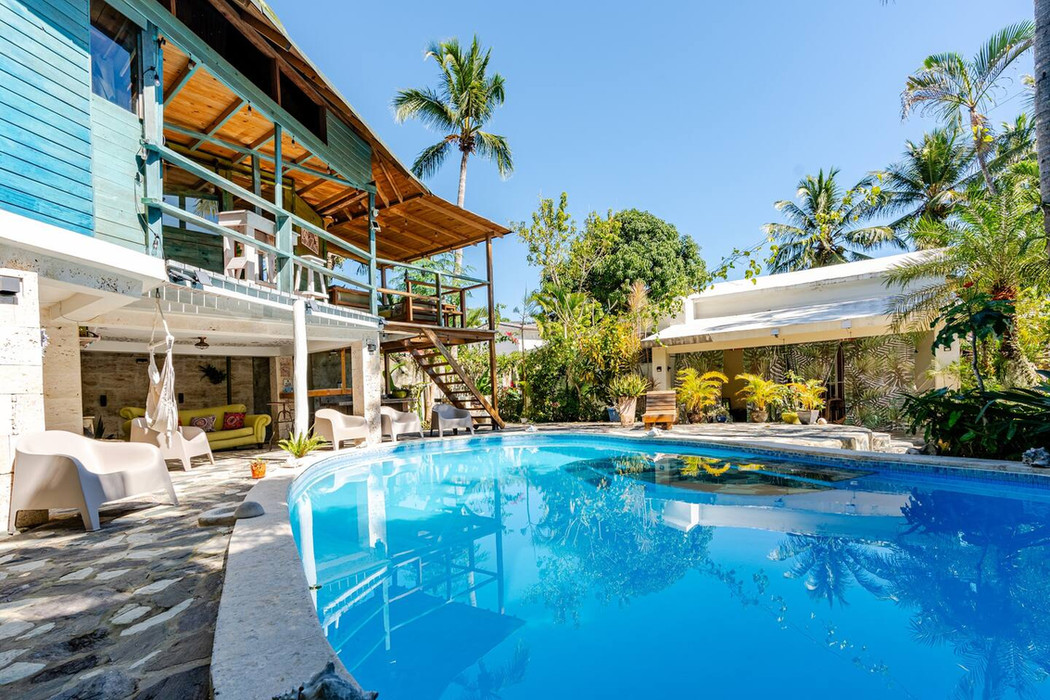 Blue swimming pool in front of a tropical house building with palm trees.