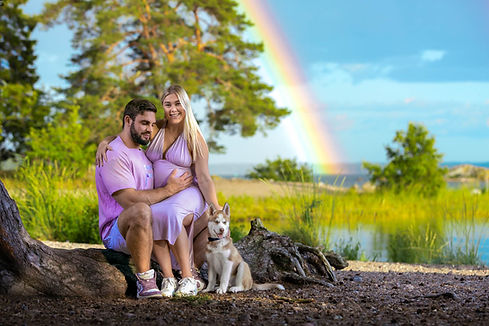 Mies ja naien istuvat koiransa kanssa puun juurella merenrannalla ja takana näkyy sateenkaari