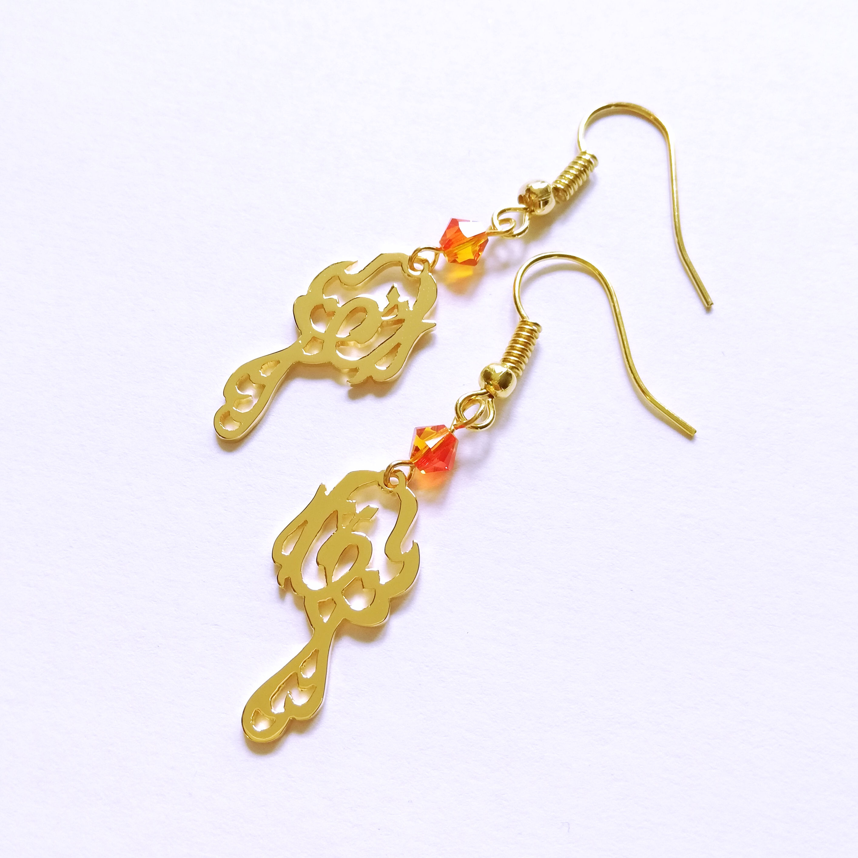 Gold phoenix earring with orange Swarovski beads on top laying on white. The bird shape is designed with Arabic.