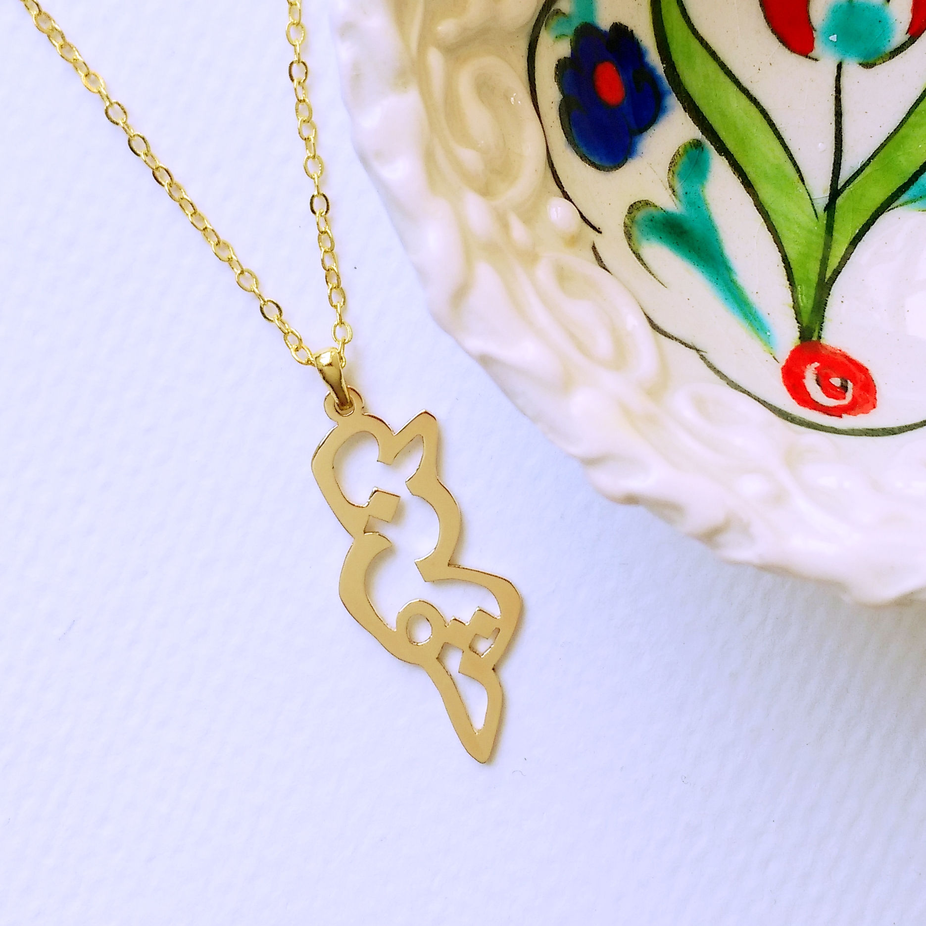 Gold Tunisia country shape pendant designed with Arabic. Lying on white next to Turkish pottery.