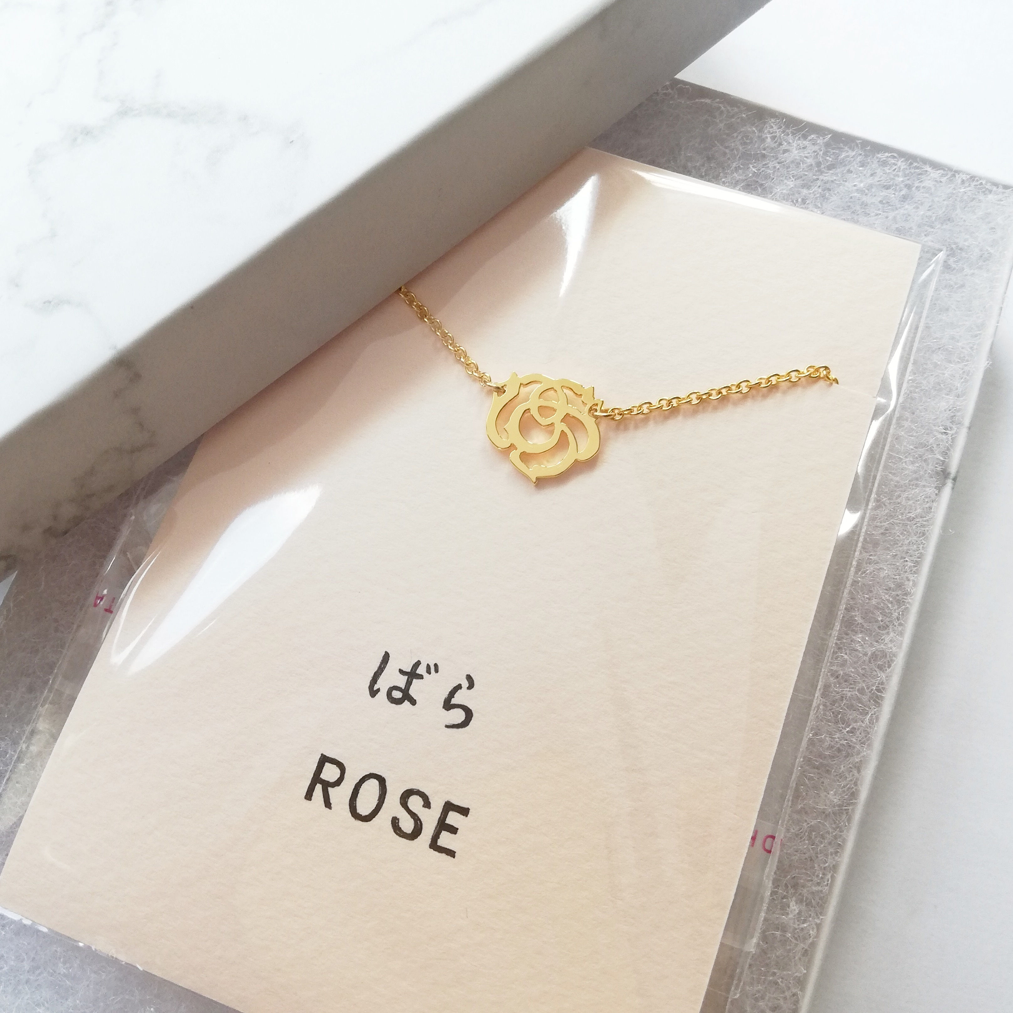 Gold rose flower necklace designed with Japanese word is set on a card, resting in cardboard box.