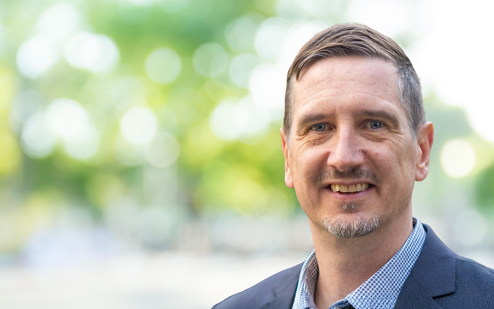 Smiling man (Emmanuel Kuntsche) in a blue suit and checkered shirt stands outdoors with a blurred green and yellow background, suggesting a sunny day.