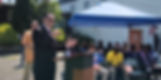 Rick Koreyva speaks at podium outdoors under a blue canopy.