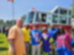 Group of six people smiling in front of a fire truck, wearing colorful attire. A Liberian flag and sunny outdoor setting are visible.