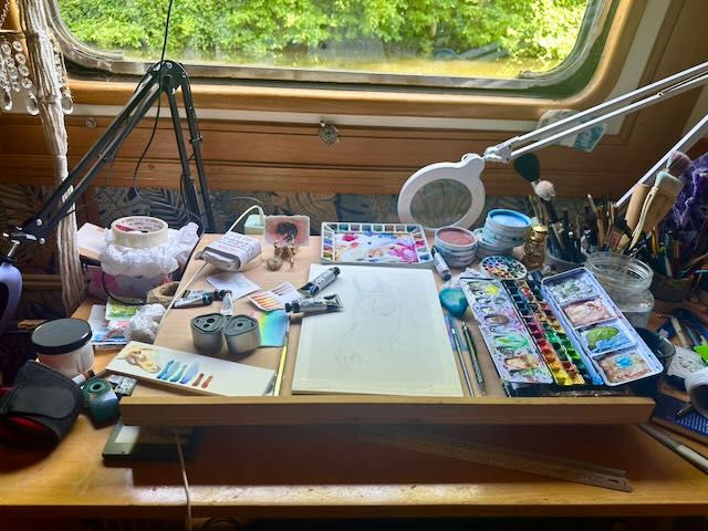 a desk covered in art equipment including a sketch of a horse, paints, paint brushes, and palettes.