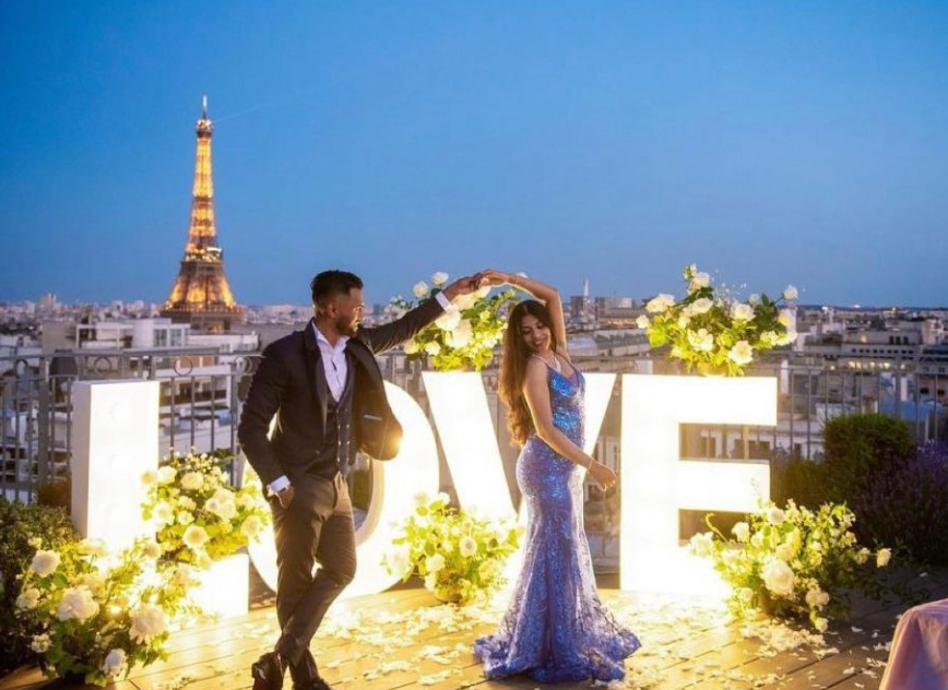 Couple dances near illuminated "LOVE" sign, surrounded by flowers, with Eiffel Tower in the background. Evening sky, romantic mood. Paris Terrace Proposal. Paris Christmas Proposal.