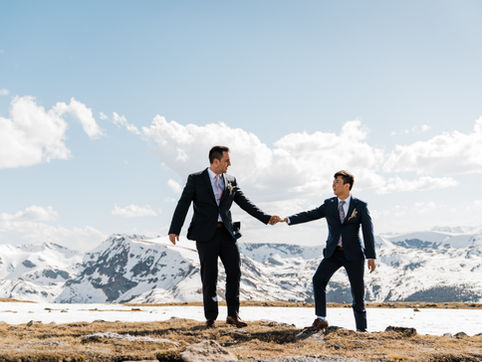 A gay couple holding hands on a snowy mountain ridge with a dramatic blue sky backdrop.