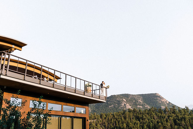 A breathtaking panoramic view of a Colorado mountain range at sunset, featuring a bride and groom standing on a wooden balcony overlooking the rolling foothills and distant peaks during an intimate mountain wedding.