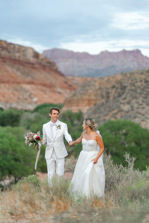 Bride and groom walking hand-in-hand through the desert brush with a distant mountain view at their Zion National Park wedding.