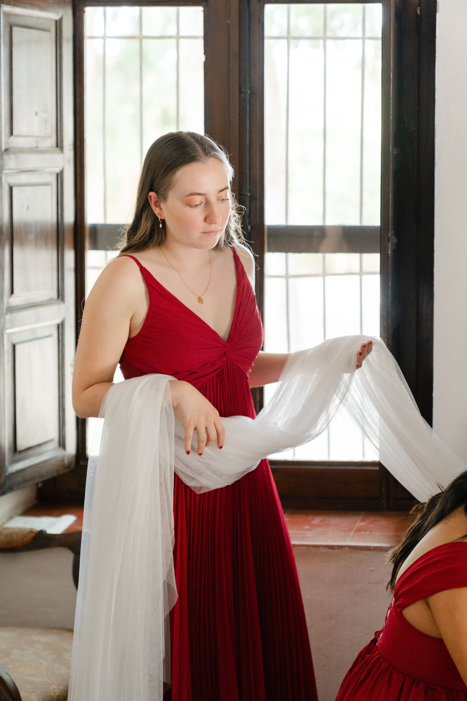 Bridesmaid in a red gown holding a white bridal veil in front of a large Mediterranean-style window.