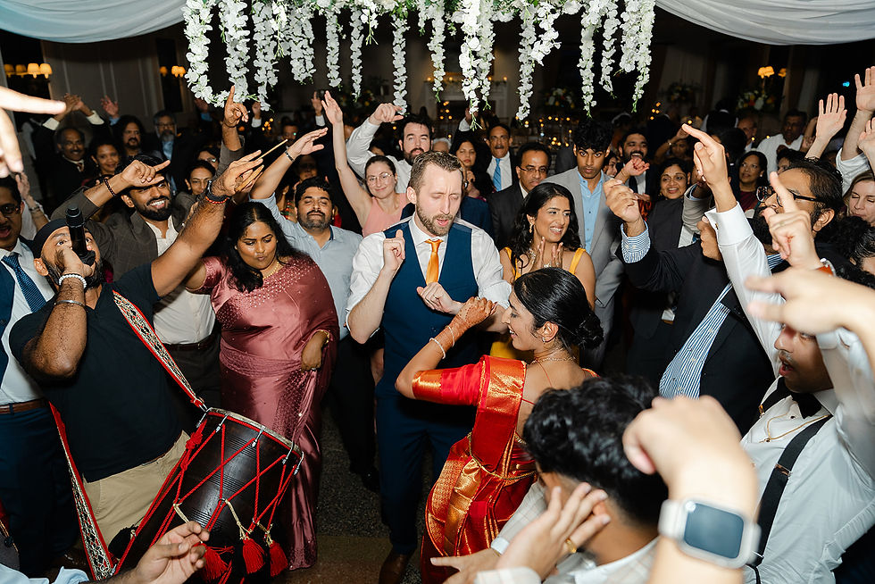 A lively wedding party with guests dancing joyfully; vibrant sarees and suits; a drummer energizes the crowd beneath floral decorations.