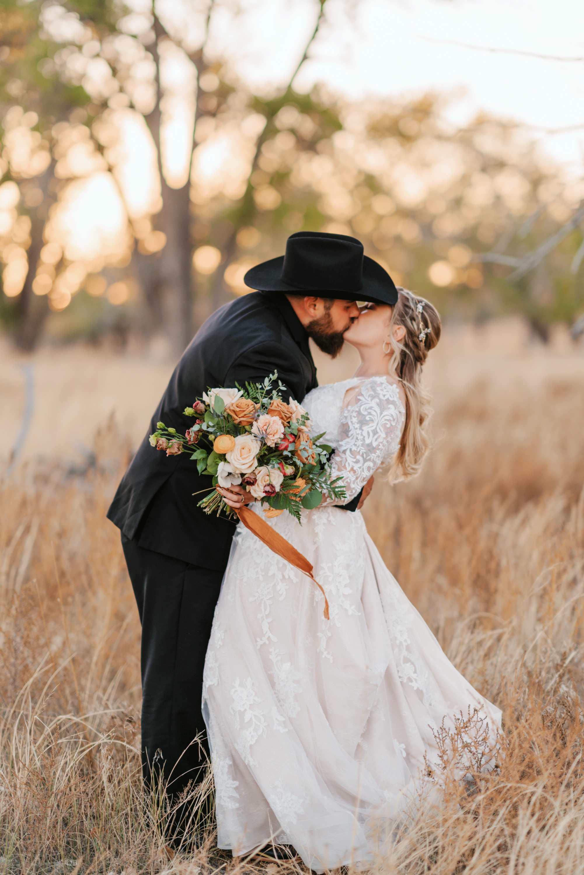 A bride in a lace wedding dress and a groom in a black hat sharing a kiss in a rustic field at sunset.