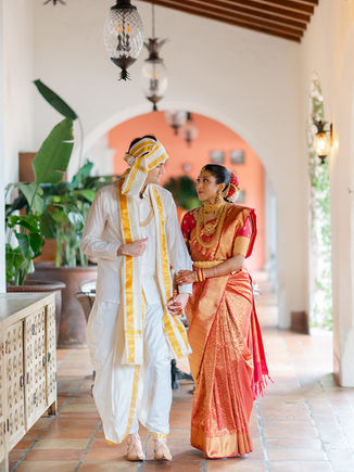A bride and groom in ornate traditional attire walking through an arched hallway decorated with floral arrangements.