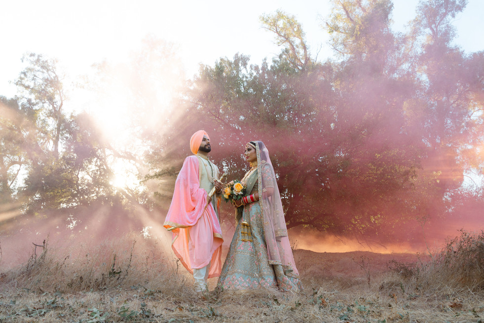 An ethereal, wide-angle portrait of a South Asian bride and groom standing in a sun-drenched field in Sacramento, CA, showcasing a luxury peach turban and gold bridal lehenga against a hazy sunset.