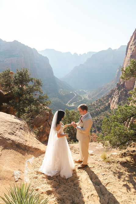 A bride and groom standing together at the edge of a dramatic sandstone cliff at Canyon Overlook in Zion National Park.
