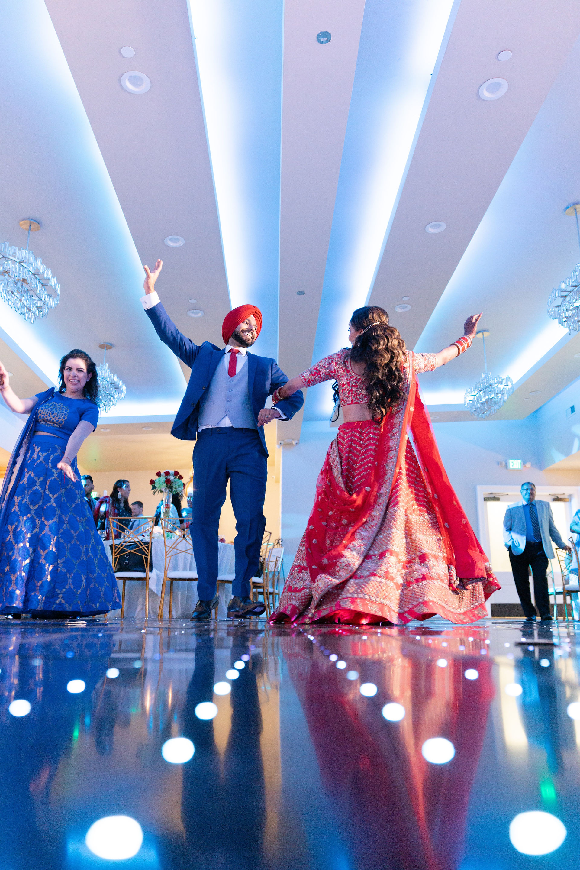 A high-energy South Asian wedding reception at White Oak reception hall in Sacramento, featuring the bride in a vibrant red lehenga and the groom in a blue suit on a glowing LED dance floor.