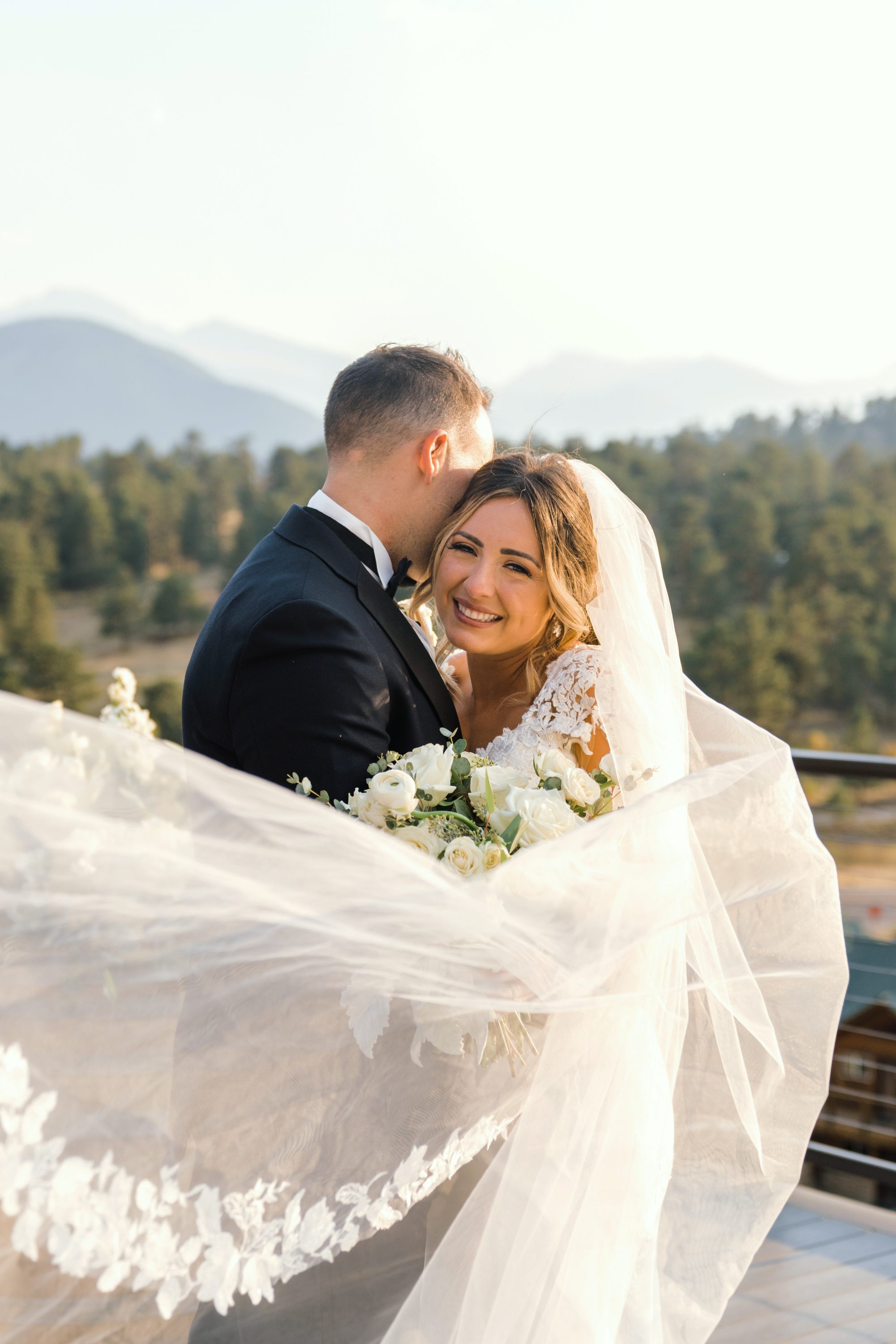 A groom whispering to his bride as her long white veil catches the wind against a scenic Rocky Mountain National Park backdrop in SkyView at Fall River Village.