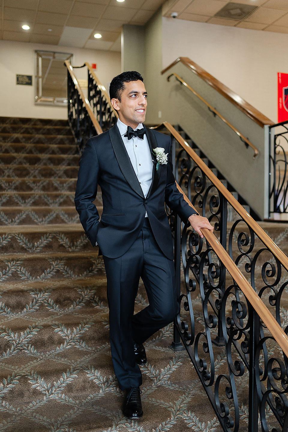 Portrait of groom Vince in a classic black tuxedo smiling on a grand staircase at the Clayton Plaza Hotel during his St. Louis wedding festivities.