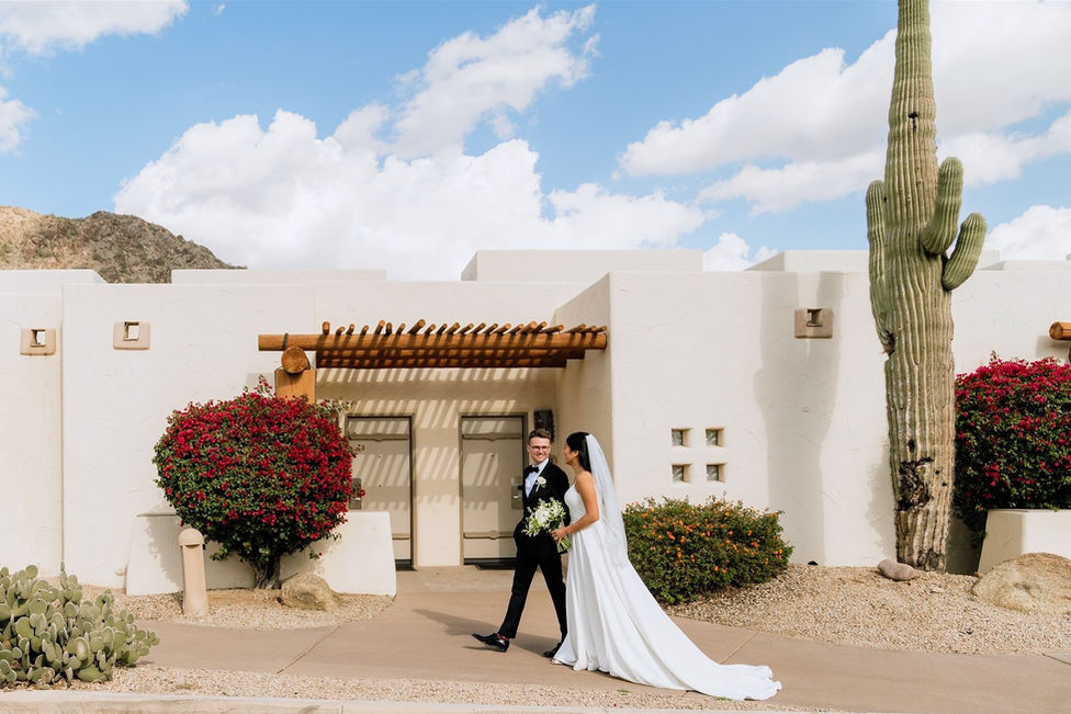 A bride and groom sharing a romantic moment in front of the white Adobe-style architecture of a luxury Arizona resort, framed by vibrant desert landscaping.