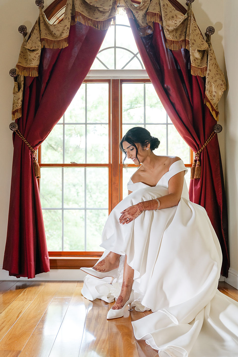Bride Sarah in her classic white wedding dress sitting by a window with red drapery during her morning prep at her family home in St. Louis.
