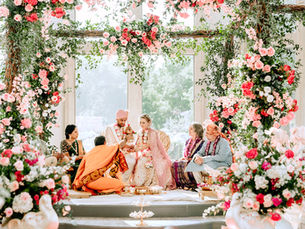 A couple in traditional attire sit among family at a floral-adorned ceremony. Pink roses and greenery create a vibrant, festive atmosphere.