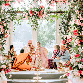 A couple in traditional attire sit among family at a floral-adorned ceremony. Pink roses and greenery create a vibrant, festive atmosphere.
