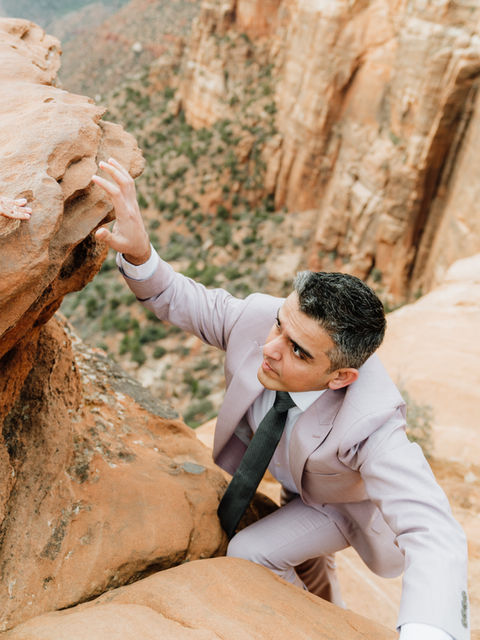 A groom in a grey suit and tie leaning against a unique red rock formation during an adventure elopement photoshoot.