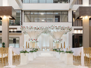Elegant indoor wedding setup with gold chairs, white drapes, and floral arches. Red and white flowers adorn the venue, creating a classy ambiance.
