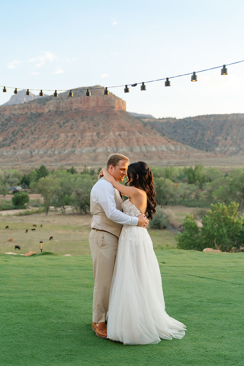 The bride and groom sharing a quiet embrace on a green lawn in Rockville, Utah, during their adventure wedding in Zion National Park, with red rock mesas in the background.