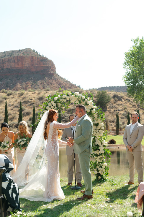 A stunning wide-angle view of the wedding ceremony by the pond, featuring the couple at the altar with the desert cliffs of Gooseberry Mesa in the background.