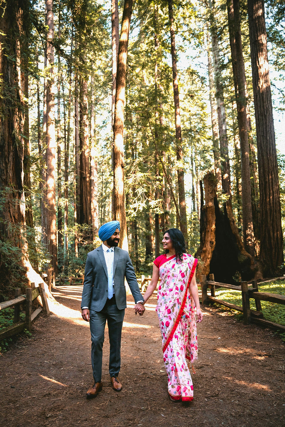 A groom in a grey suit and blue turban holding hands with a bride in a red and white floral sari while walking through a redwood forest.